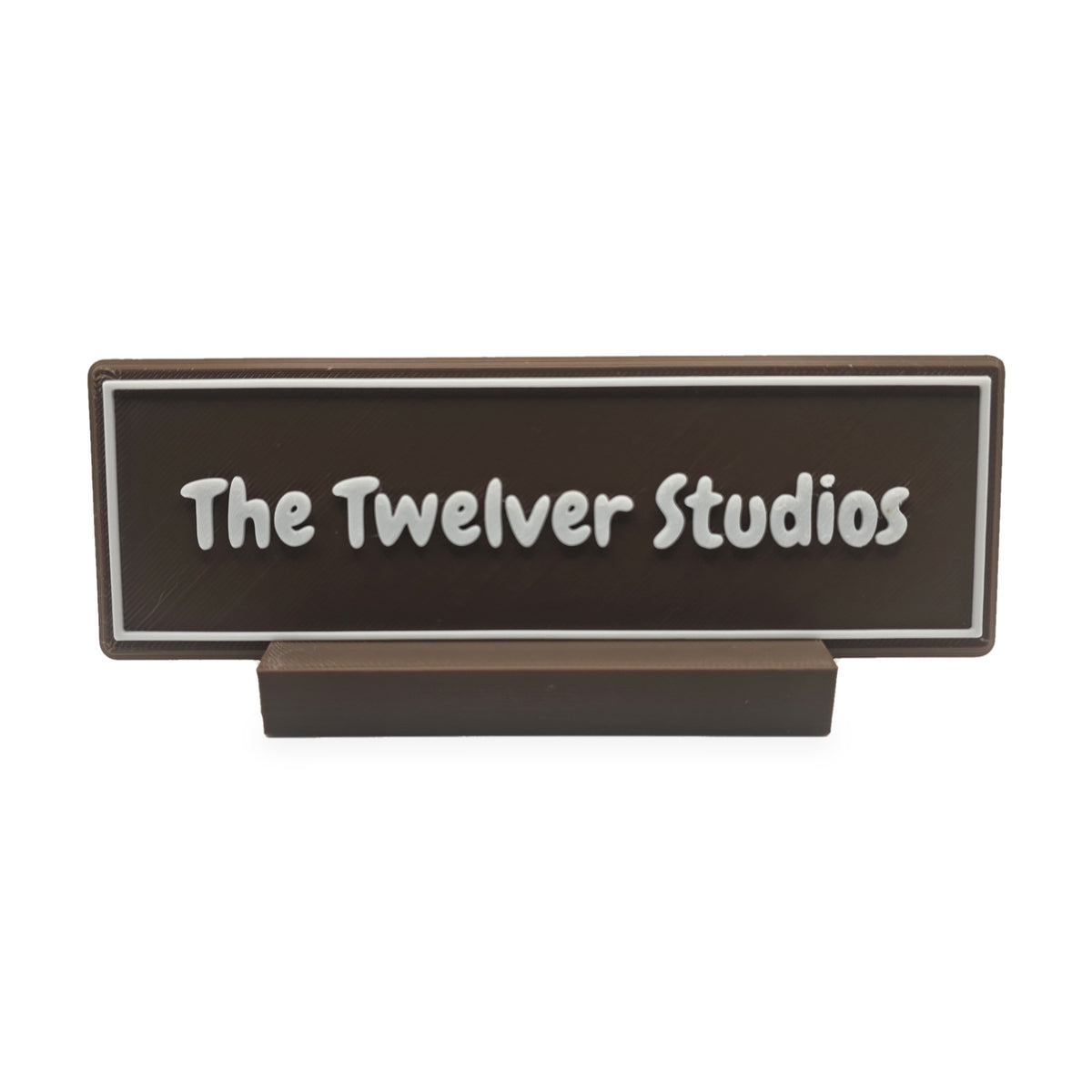 Small brown desk name plate in its brown slotted foot, with white raised lettering and border reading The Twelver Studios