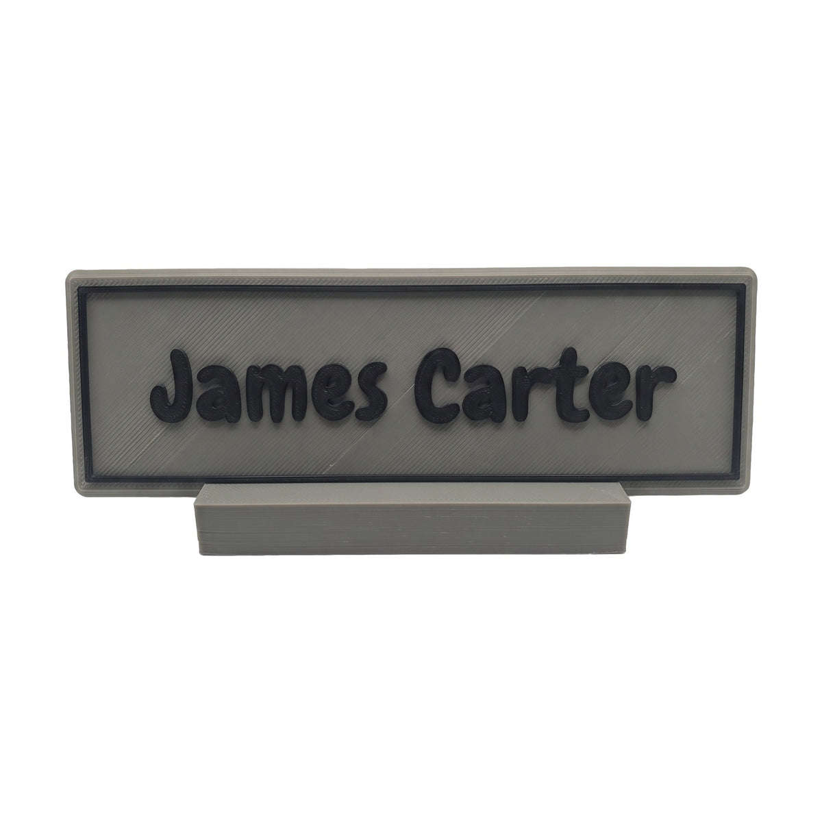 Example personalisation: Small grey desk name plate and foot with black raised lettering and border reading James Carter
