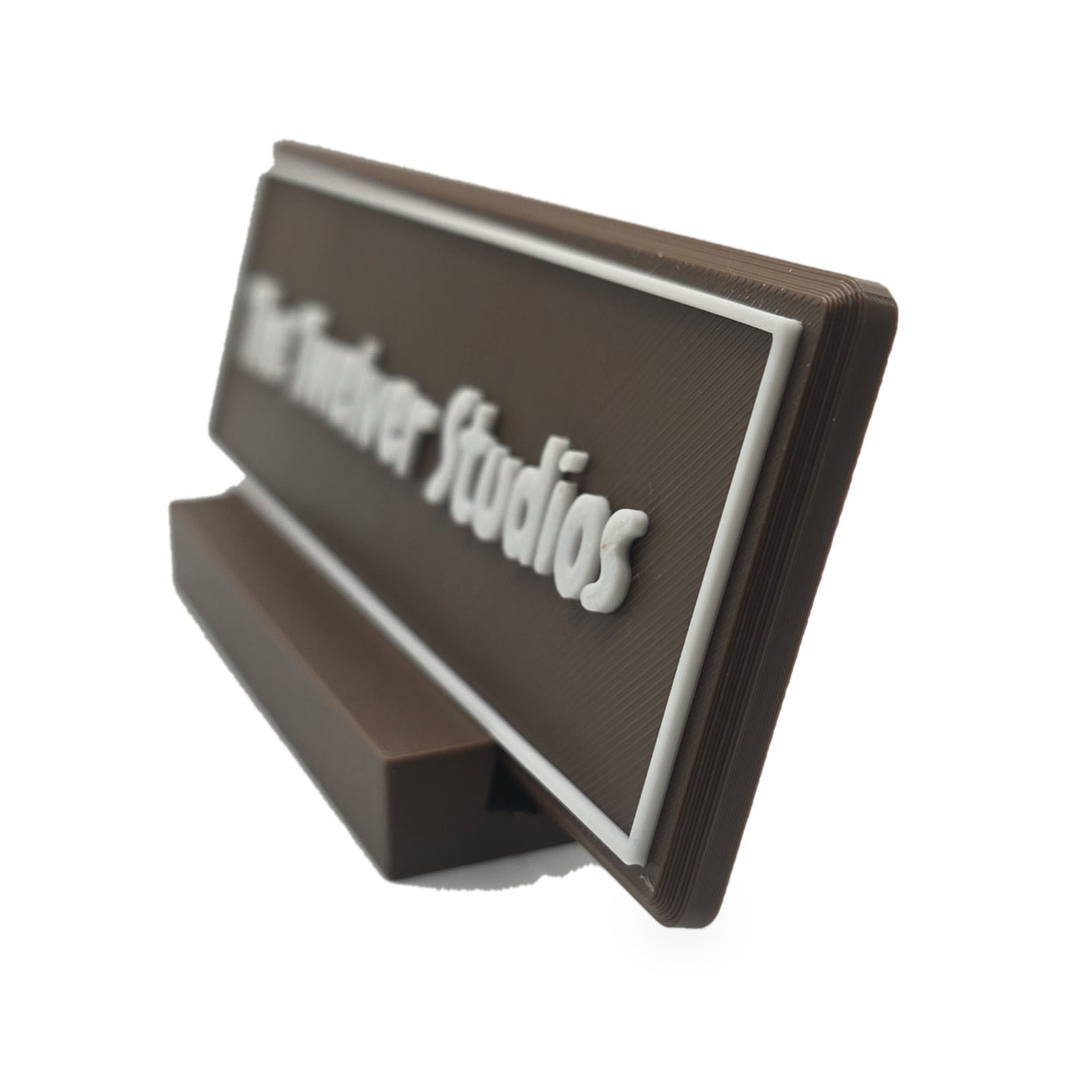 Brown desk name plate with white raised lettering and border reading The Twelver Studios, seen from the side in its brown slotted foot